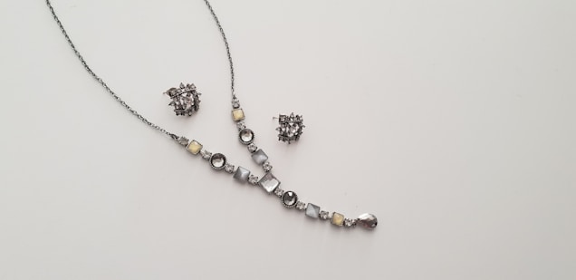 A delicate and elegant jewelry set consisting of a necklace and a pair of matching earrings is displayed against a plain light background. The necklace features a series of round and square gemstones in shades of silver and light yellow, arranged along a thin chain. The earrings are intricate clusters of similarly colored stones.