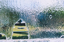 A sheet of glass features a jagged hole with a distinct outline, surrounded by water droplets and a textured surface. Through the opening, a blurred view of a grassy area and distant objects is visible, giving a sense of depth and contrast between the clear and opaque portions.