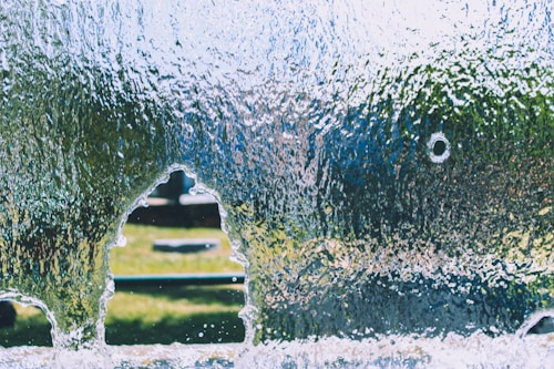 A sheet of glass features a jagged hole with a distinct outline, surrounded by water droplets and a textured surface. Through the opening, a blurred view of a grassy area and distant objects is visible, giving a sense of depth and contrast between the clear and opaque portions.