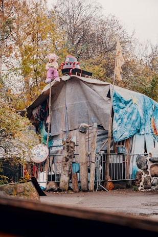 A makeshift, artistic shelter surrounded by trees in an autumn environment. The structure is covered with tarps and decorated with artwork, including a prominent painted star on a blue background. On top of the shelter, a large teddy bear dressed in pink and a whimsical hat sits alongside a playful prop with a painted face. Various pieces of wood and artistic elements surround the space, creating an eclectic and whimsical atmosphere.