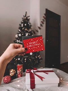 A hand is holding a festive red card with the text 'Merry Christmas' in front of a decorated Christmas tree. The tree is adorned with glowing lights and ornaments. In the foreground, there is a white gift box tied with a red ribbon, accompanied by holiday-themed items like a decorative ball and a candle with Christmas motifs. The setting appears cozy and warm, filled with holiday cheer.