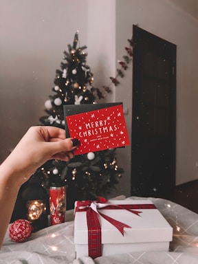 A hand is holding a festive red card with the text 'Merry Christmas' in front of a decorated Christmas tree. The tree is adorned with glowing lights and ornaments. In the foreground, there is a white gift box tied with a red ribbon, accompanied by holiday-themed items like a decorative ball and a candle with Christmas motifs. The setting appears cozy and warm, filled with holiday cheer.
