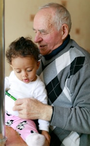 An elderly man is holding a young child on his lap. The child is wearing a white shirt and pink pants featuring a cartoon character pattern. The man is wearing a gray sweater with a black and white argyle pattern and appears to be showing the child something in his hand.
