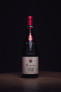 A wine bottle with a red cap and an elegant label displaying the name 'Rasteau'. The bottle stands on a wooden surface against a dark background, highlighting its sophisticated design.