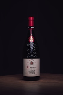 A wine bottle with a red cap and an elegant label displaying the name 'Rasteau'. The bottle stands on a wooden surface against a dark background, highlighting its sophisticated design.