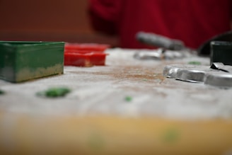 The foreground features a dusting of flour on a surface with various cookie cutters. There are green and silver rectangular and circular shapes, suggesting a baking activity in progress. Red tones in the background hint at a festive or holiday atmosphere.