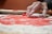 A hand with flour-covered fingers presses a circular cookie cutter onto a sheet of red dough, which is sprinkled with flour. A plate remains visible in the background, along with other baking items.