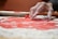 A hand with flour-covered fingers presses a circular cookie cutter onto a sheet of red dough, which is sprinkled with flour. A plate remains visible in the background, along with other baking items.