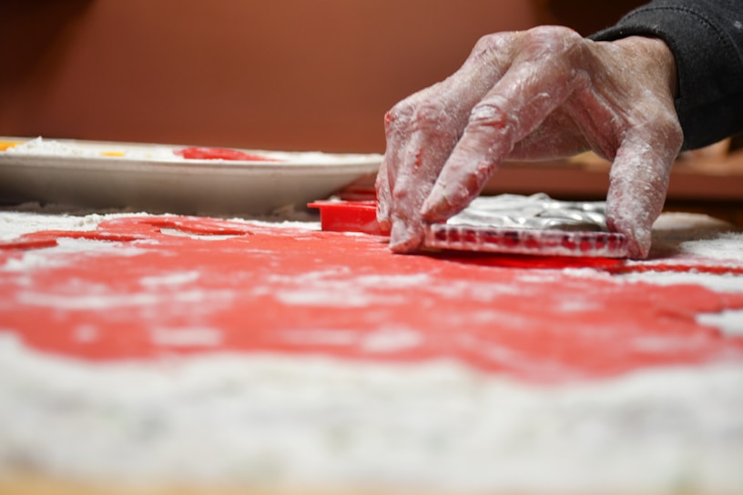 A hand with flour-covered fingers presses a circular cookie cutter onto a sheet of red dough, which is sprinkled with flour. A plate remains visible in the background, along with other baking items.