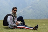 A person wearing sunglasses and a scarf is sitting on a grassy field with mountains in the background. They are holding a camera with a zoom lens and have a backpack beside them. The scene suggests an outdoor adventure or photography trip.