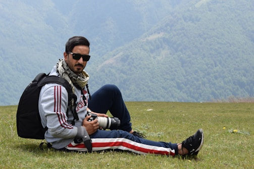 A person wearing sunglasses and a scarf is sitting on a grassy field with mountains in the background. They are holding a camera with a zoom lens and have a backpack beside them. The scene suggests an outdoor adventure or photography trip.