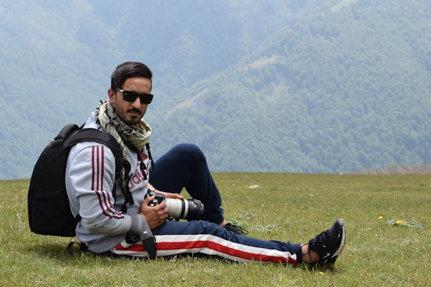 A person wearing sunglasses and a scarf is sitting on a grassy field with mountains in the background. They are holding a camera with a zoom lens and have a backpack beside them. The scene suggests an outdoor adventure or photography trip.