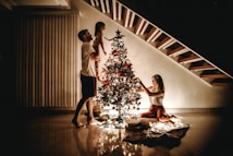 A family decorates a Christmas tree placed under a staircase. The warm light from the tree illuminates the room, highlighting the joyful atmosphere. A man holds a child up to place an ornament, while a woman kneels to adjust decorations.