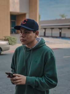 A person wearing a green hoodie and a black cap with a red letter C on it is holding a smartphone. The setting appears to be outdoors in a parking lot, with a car and some buildings visible in the background.