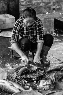 A woman wearing a plaid shirt is sitting on a small stool, using a large knife to clean or butcher fish on a wet, textured surface. The surrounding area is messy with fish scales and water, and multiple fish are visible around her. She is focused on her task, and there is a sense of concentration in her posture.