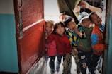 A group of children stand at an open doorway, peering inside with curious expressions. They are dressed in colorful clothing, with a mix of sweaters and jackets. The setting appears intimate and friendly, suggesting a sense of community or playfulness.