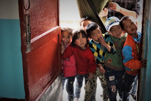 A group of children stand at an open doorway, peering inside with curious expressions. They are dressed in colorful clothing, with a mix of sweaters and jackets. The setting appears intimate and friendly, suggesting a sense of community or playfulness.