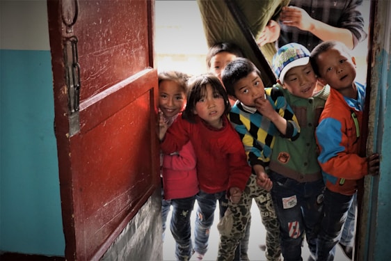 A group of children stand at an open doorway, peering inside with curious expressions. They are dressed in colorful clothing, with a mix of sweaters and jackets. The setting appears intimate and friendly, suggesting a sense of community or playfulness.