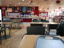 A spacious cafeteria with wooden flooring, featuring several empty tables and chairs. The counter area at the back displays a variety of food and beverage options, with menus hanging above. The decor includes red and white hues, with circular hanging decorations.