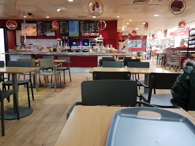 A spacious cafeteria with wooden flooring, featuring several empty tables and chairs. The counter area at the back displays a variety of food and beverage options, with menus hanging above. The decor includes red and white hues, with circular hanging decorations.