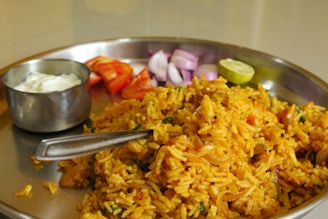 A stainless steel plate containing a serving of spicy yellow rice garnished with herbs. Sides include sliced red tomatoes, chopped purple onions, a small wedge of lime, and a small bowl of white yogurt.