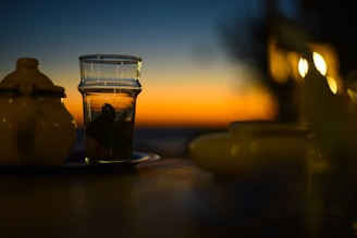 A glass of tea with mint leaves sits on a table next to a ceramic teapot, with a beautiful sunset in the background. The sky shifts from deep blue to orange, creating a calming and picturesque atmosphere. The lighting is warm and soft, with blurred lights in the distance adding to the serene ambiance.