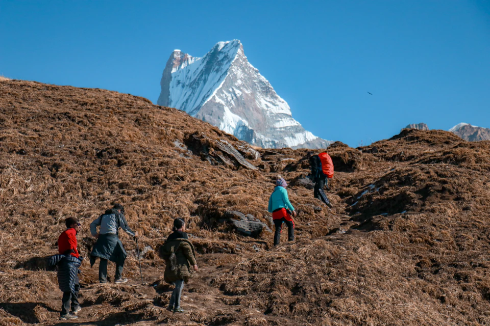 The image depicts a group of four hikers ascending a mountain trail. The hikers are wearing backpacks and moving towards the right side of the frame, with one hiker slightly ahead of the others. In the background, a majestic snow-capped peak looms over the scene, adding to the sense of adventure and grandeur.