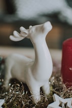 A white ceramic deer figurine with antlers is placed on a decorative surface, surrounded by gold tinsel and a large red candle. Soft and blurred elements in the background suggest a cozy indoor setting.