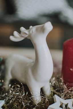 A white ceramic deer figurine with antlers is placed on a decorative surface, surrounded by gold tinsel and a large red candle. Soft and blurred elements in the background suggest a cozy indoor setting.