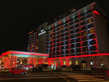 A well-lit hotel facade at night, featuring colorful red and white lighting. Several cars are parked in front, and there are decorative lights wrapped around the entrance columns. The bold, illuminated sign displays the name of the hotel.