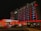 A well-lit hotel facade at night, featuring colorful red and white lighting. Several cars are parked in front, and there are decorative lights wrapped around the entrance columns. The bold, illuminated sign displays the name of the hotel.