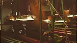 A view of a weaving loom in a textile factory. The machinery is predominantly green with intricate parts and threads running through it. The ambient light casts a warm glow, highlighting the complexity of the weaving process.