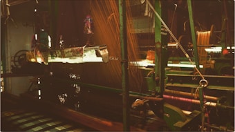A view of a weaving loom in a textile factory. The machinery is predominantly green with intricate parts and threads running through it. The ambient light casts a warm glow, highlighting the complexity of the weaving process.