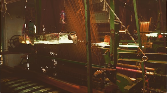 A view of a weaving loom in a textile factory. The machinery is predominantly green with intricate parts and threads running through it. The ambient light casts a warm glow, highlighting the complexity of the weaving process.