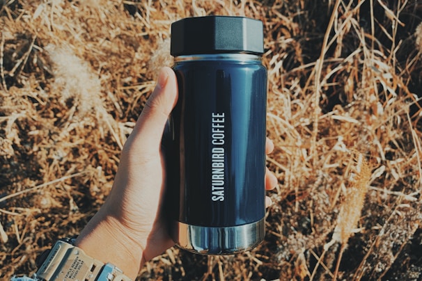 A hand holds a dark, cylindrical coffee thermos labeled 'SATURNBIRD COFFEE' against a background of dry grass or wheat. The scene is brightly lit, suggesting a sunny day.