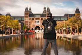 A person stands by a reflective water feature, capturing a photograph of a large, architecturally detailed building with prominent towers. Lush trees surround the area, and the iconic red and white 'I Amsterdam' sign is visible at the base of the building.