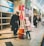A person dressed as a bell-ringing Salvation Army worker stands next to a young child wearing a pink coat who is placing something into an orange donation bucket. They are situated in a busy indoor area, possibly a shopping mall, with other people visible in the background.