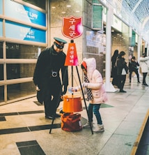 A person dressed as a bell-ringing Salvation Army worker stands next to a young child wearing a pink coat who is placing something into an orange donation bucket. They are situated in a busy indoor area, possibly a shopping mall, with other people visible in the background.