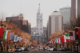 A bustling city street lined with international flags, converging towards a grand, historic building with a prominent clock tower. Tall modern skyscrapers and buildings frame the background, while cars and pedestrians are visible in the foreground.