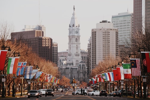 A bustling city street lined with international flags, converging towards a grand, historic building with a prominent clock tower. Tall modern skyscrapers and buildings frame the background, while cars and pedestrians are visible in the foreground.