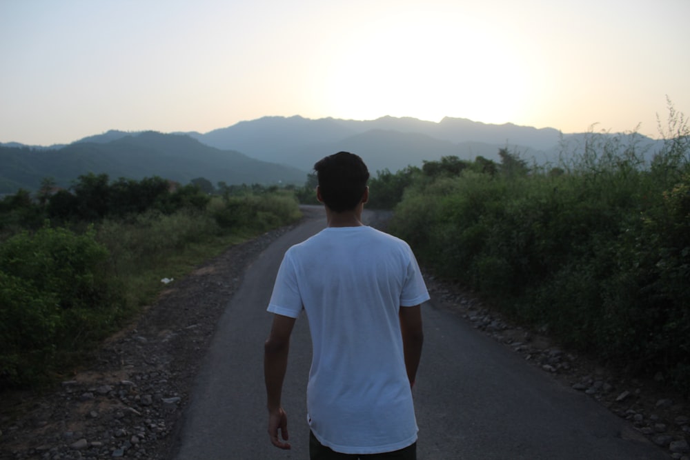 man wearing white shirt walking on road surrounded by bushes