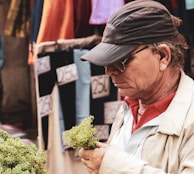 An older man in a market setting is examining a bundle of what appears to be herbs or greens. He is wearing a cap and glasses, dressed in a beige jacket over a red collared shirt. In the background, there are various clothing items on display with price tags visible.