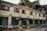 A charming, two-story building with wooden shutters on the windows and an exterior decorated in earthy tones, housing a cafe named 'Fika' complete with decorative signage. The cafe has several dark brown awnings and seating arrangements with chairs and tables outside. Green plants in pots line the front, adding a touch of nature. Overhead, the sky is partly cloudy, and the background features lush trees.