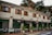 A charming, two-story building with wooden shutters on the windows and an exterior decorated in earthy tones, housing a cafe named 'Fika' complete with decorative signage. The cafe has several dark brown awnings and seating arrangements with chairs and tables outside. Green plants in pots line the front, adding a touch of nature. Overhead, the sky is partly cloudy, and the background features lush trees.