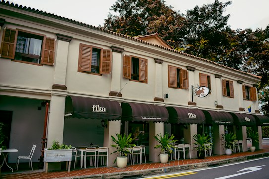 A charming, two-story building with wooden shutters on the windows and an exterior decorated in earthy tones, housing a cafe named 'Fika' complete with decorative signage. The cafe has several dark brown awnings and seating arrangements with chairs and tables outside. Green plants in pots line the front, adding a touch of nature. Overhead, the sky is partly cloudy, and the background features lush trees.