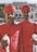 Two men are dressed in traditional red attire with matching turbans, holding silver-topped staffs. They stand in front of an ornate building with arched windows and a decorative facade. A group of people is visible in the background near the entrance. Trees flank the building, adding greenery to the scene.