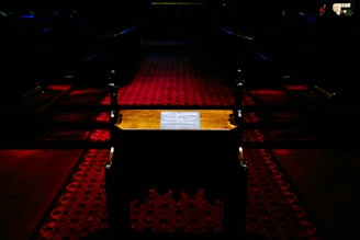 A dimly lit interior with a dark red patterned carpet in a narrow aisle. There is a small wooden table or stand in the center showcasing a sign with text, possibly indicating WiFi information. The scene has a somber and quiet atmosphere, with shadows and minimal illumination.