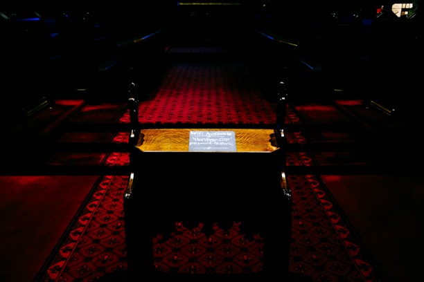 A dimly lit interior with a dark red patterned carpet in a narrow aisle. There is a small wooden table or stand in the center showcasing a sign with text, possibly indicating WiFi information. The scene has a somber and quiet atmosphere, with shadows and minimal illumination.