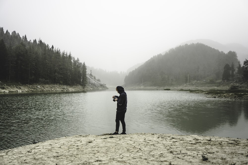 person in hoodie holds camera and stands beside body of water