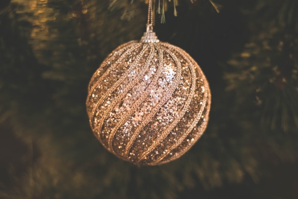 A glittering, gold and silver Christmas ornament hangs amid dark green pine tree branches. The ornament features a swirling pattern of sparkling sequins and beads, creating a festive and elegant look.